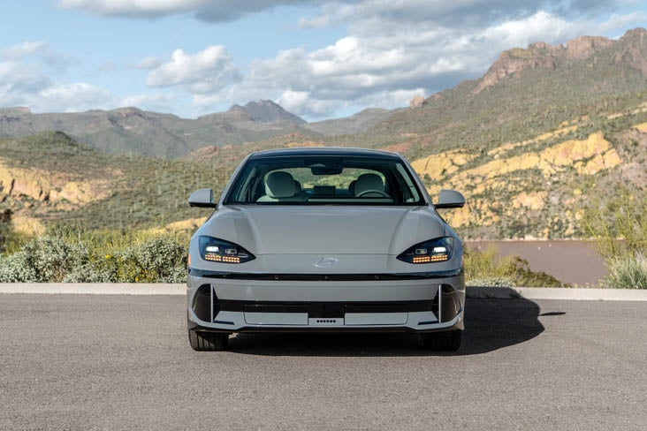 A light gray Hyundai Ioniq 6 electric car parked head-on on an asphalt road, with mountains and sparse vegetation in the background under a partly cloudy sky.