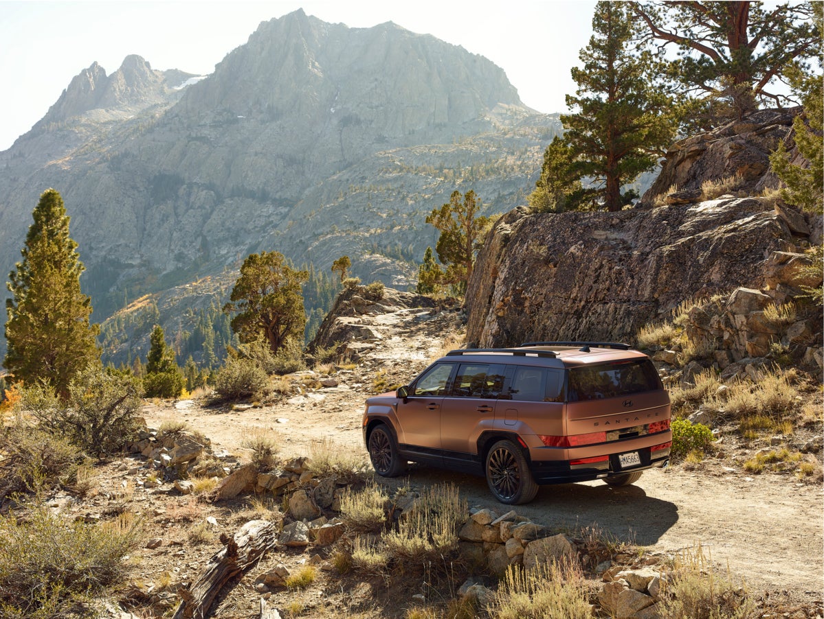 A brown 2026 Hyundai Santa Fe Hybrid in Avondale drives away from the viewer to the left on a rocky trail on the side of the face of a mountain overlooking an arid valley.